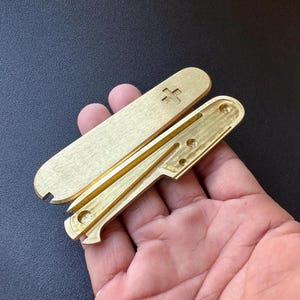 Könnte beinhalten: Ein Messing-Multitool mit einem Pluszeichen auf dem oberen Teil. Das Werkzeug ist geöffnet und zeigt das Innendesign. Das Werkzeug hat ein schlankes, minimalistisches Design und scheint aus massivem Messing zu bestehen. Das Werkzeug wird in einer Hand gehalten.