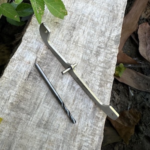 Puede incluir: Una herramienta de metal plateado con un extremo curvo y una broca recta y delgada. La herramienta está sobre una superficie de madera desgastada, con hojas verdes y follaje marrón en el fondo. La broca es de color gris oscuro.