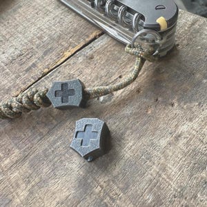 Op de afbeelding: Close-up van een multitool met een koord en twee grijze metalen Zwitserse kruis kralen. De kralen liggen op een houten oppervlak. Het koord heeft een camouflagepatroon.