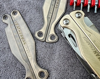 Könnte beinhalten: Zwei goldfarbene Leatherman Charge Multitools mit Messingakzenten. Die Werkzeuge sind geöffnet und zeigen die Klingen und andere Werkzeuge. Die Werkzeuge befinden sich auf einer grauen, strukturierten Oberfläche. Der Text "LEATHERMAN CHARGE" und "BRASS CU" ist in die Werkzeuge graviert.