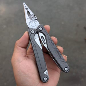 Op de afbeelding: Een zwarte Leatherman Charge multitool met titanium handvatten. De tool is open met de tangen uitgeschoven. De tool heeft een verscheidenheid aan andere tools, waaronder een mes, een zaag en een schroevendraaier.