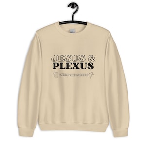 Può includere: Una felpa color crema con le parole "JESUS & PLEXUS" in nero, insieme alla frase "KEEP ME GOING" e una piccola grafica a forma di croce e tazza di caffè. La felpa è appesa a una gruccia nera.