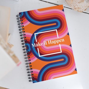 May include: Spiral-bound notebook with a retro design. The cover features a vibrant pattern of curved lines in orange, pink, blue, and red. The text "Make it Happen" is displayed in a white font within a white square. The notebook is on a white surface.