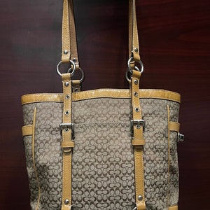 May include: A brown and tan Coach tote bag with a signature pattern and leather accents. The bag has two top handles and a buckle closure.