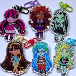Peut inclure: Porte-clés en acrylique avec des illustrations de dessins animés de divers personnages. Chaque porte-clés a un design de personnage différent avec des couleurs vives et est attaché avec un fermoir en métal. Les porte-clés sont conçus dans un style de dessin animé.