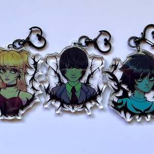 Peut inclure: Trois porte-clés en acrylique avec des portraits de style anime. Chaque porte-clés a un fermoir noir en forme de cœur et un bord clair et irrégulier avec un motif noir en forme de vigne. Les portraits représentent des personnages avec des cheveux et des vêtements distinctifs.