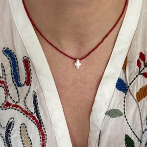 May include: A delicate red beaded necklace with a small, white, cross-shaped pendant. The necklace is worn over a white blouse with colorful embroidered floral and abstract designs.