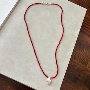 May include: A red beaded necklace with a white, irregular-shaped pendant. The necklace has a silver clasp and is displayed on a light gray surface. The beads are small and evenly spaced, creating a delicate look.