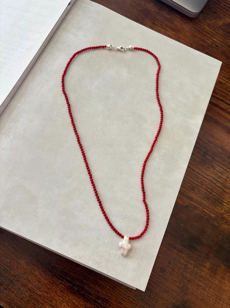 May include: A red beaded necklace with a white, irregular-shaped pendant. The necklace has a silver clasp and is displayed on a light gray surface. The beads are small and evenly spaced, creating a delicate look.