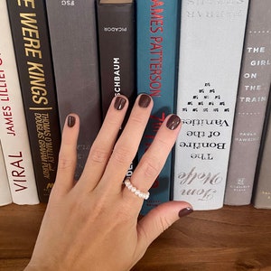 May include: A hand wearing a pearl ring with a row of white pearls. The hand is resting on a bookshelf with several books, including "We Were Kings" by Thomas O'Malley and Douglas Graham Purdy, "The Girl on the Train" by Paula Hawkins, and "The Bonfire of the Vanities" by Tom Wolfe.