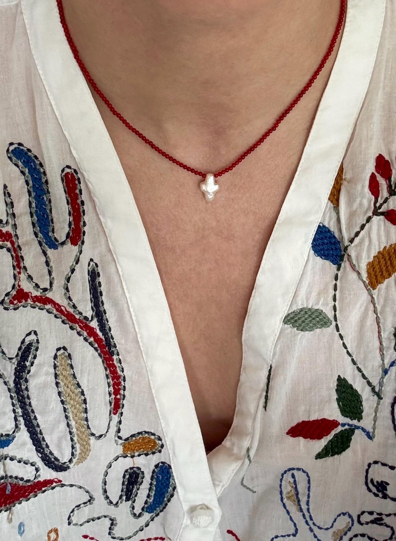 May include: A delicate red beaded necklace with a small, white, cross-shaped pendant. The necklace is worn over a white blouse with colorful embroidered floral and abstract designs.