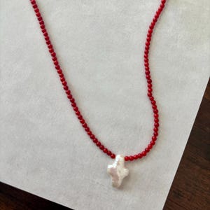 May include: A red beaded necklace with a white cross pendant. The necklace features small, round red beads strung together, forming the chain. The pendant is a white, irregularly shaped cross, adding a unique touch to the jewelry.