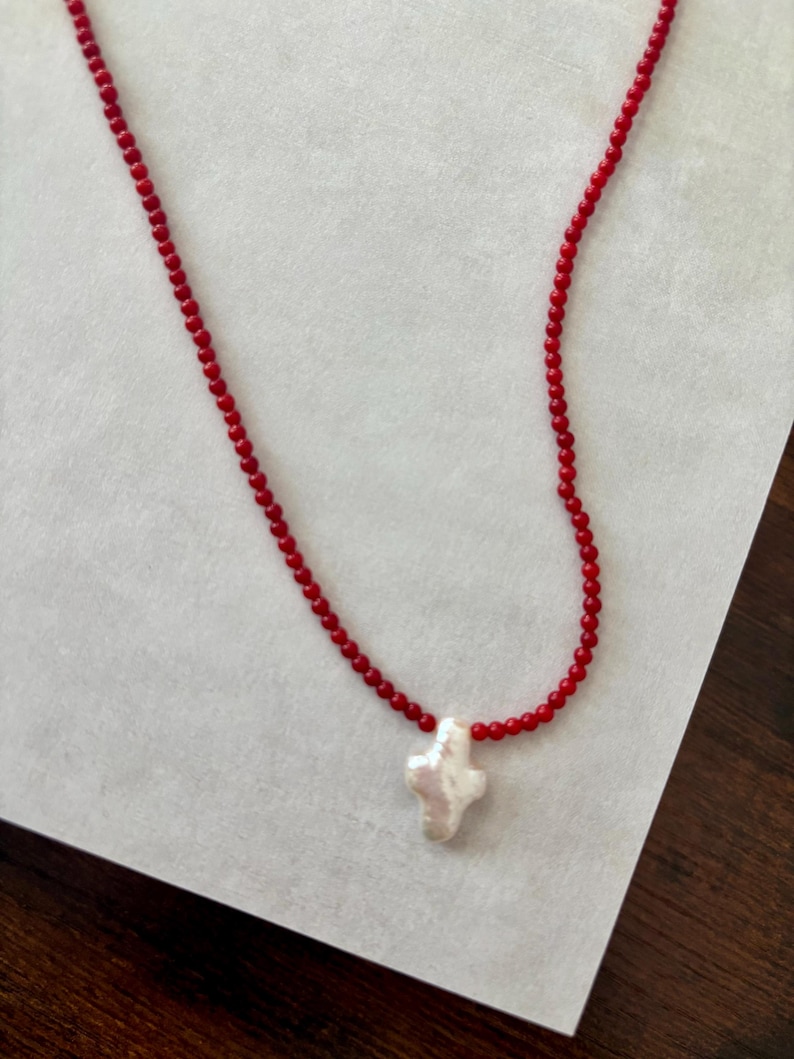 May include: A red beaded necklace with a white cross pendant. The necklace features small, round red beads strung together, forming the chain. The pendant is a white, irregularly shaped cross, adding a unique touch to the jewelry.