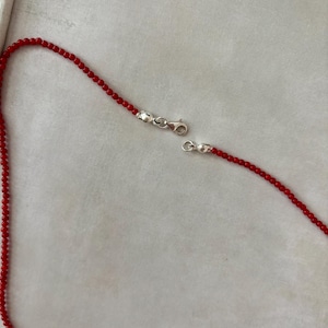May include: A red beaded necklace with a silver clasp. The necklace is composed of small, round red beads strung together. The silver clasp is open, showing the necklace's two ends.