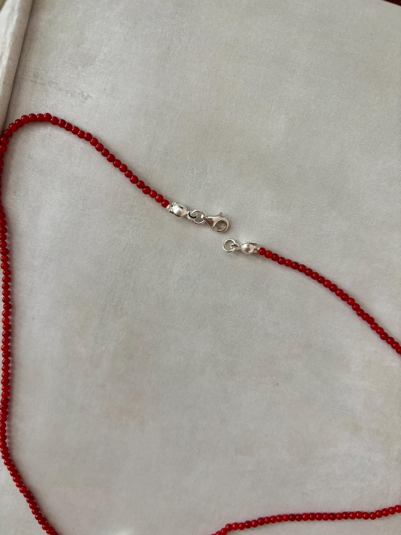 May include: A red beaded necklace with a silver clasp. The necklace is composed of small, round red beads strung together. The silver clasp is open, showing the necklace's two ends.