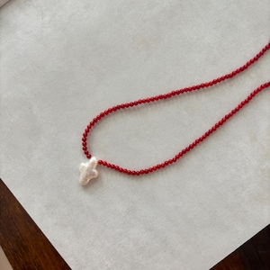 May include: A red beaded necklace with a white cross pendant. The necklace features small, round red beads strung together, with a cross-shaped pendant at the center. The cross appears to be made of a textured, off-white material.