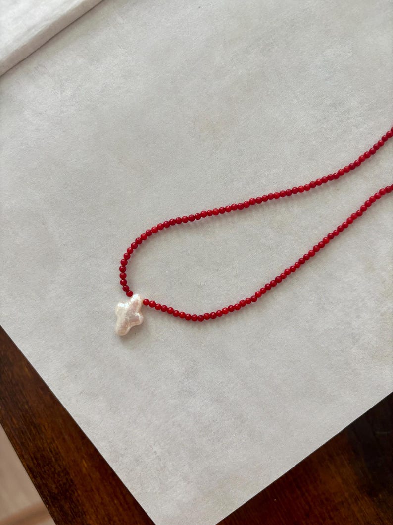 May include: A red beaded necklace with a white cross pendant. The necklace features small, round red beads strung together, with a cross-shaped pendant at the center. The cross appears to be made of a textured, off-white material.