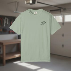 May include: A light green t-shirt with the words "Girl Dad" in black script on the front. The shirt is a solid color and has a crew neck. The background shows a garage setting.