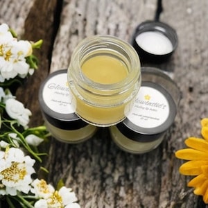 Puede incluir: Tres pequeños frascos de vidrio con tapas llenos de un bálsamo amarillo. Los frascos están etiquetados con "Flowerchild's" y "Honey & Balm".