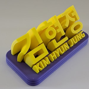 May include: A yellow 3D printed nameplate with the Korean text "김현정" and the English text "KIM HYUN JUNG" on a purple base.