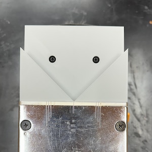 May include: A metal and plastic tool with a silver base and a white top section. The base has measurement markings and is secured with black screws. An orange and silver knob is visible on the side. The tool is held by a blue handle.