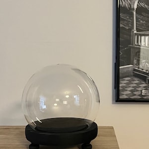 May include: A clear glass globe sits on a black, round wooden base with three small, rounded feet. The globe is empty, and the base is painted black. The setup is on a light-coloured wooden surface, with a framed black and white artwork in the background.