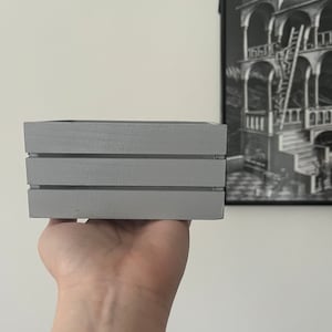 May include: A small, grey wooden box with three horizontal slats. The box is being held in a hand.