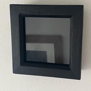 May include: A black square picture frame with a glass front. The frame is made of wood and has a simple design.