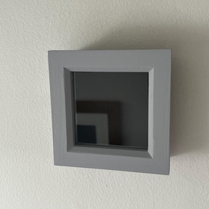 May include: A small, square, grey picture frame with a black mirror inside. The frame is hanging on a white wall.