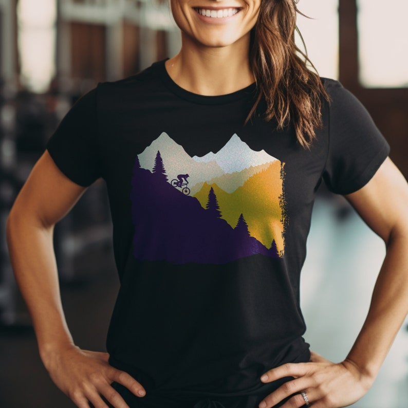 May include: Black t-shirt featuring a mountain biking graphic. The design shows a cyclist on a mountain slope with trees and mountains in the background. The color palette includes purple, green, yellow, and white.