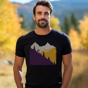 May include: Black t-shirt featuring a mountain biking graphic. The design shows a cyclist on a mountain trail with trees and layered mountain silhouettes in shades of purple, gray, and gold. The shirt is worn by a person in an outdoor setting.