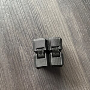 Può includere: Un giocattolo antistress nero con due sezioni quadrate che possono essere ribaltate e ruotate. Il giocattolo è in plastica e ha una finitura liscia e opaca.