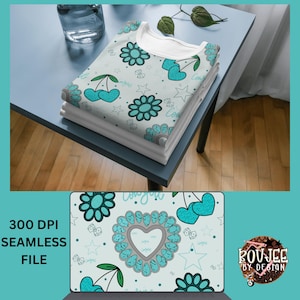 Könnte beinhalten: Ein Stapel gefalteter weißer T-Shirts mit einem hellblauen Blumen- und Kirschmuster. Das Design umfasst Blumen, Kirschen, Sterne und das Wort "Cowgirl". Die Shirts liegen auf einem blauen Tisch mit einem Glas Wasser und einer Pflanze.