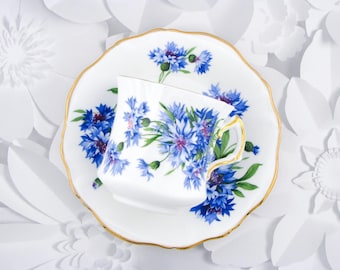 May include: A white porcelain teacup and matching saucer featuring a vibrant blue cornflower pattern with green stems and leaves. Both pieces are finished with a delicate gold trim along the scalloped edges, creating a cohesive floral design.