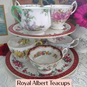 May include: A tiered serving tray displays several Royal Albert teacups and saucers. The teacups feature floral and landscape designs with gold trim. The saucers have a red border with floral patterns. The text "Royal Albert Teacups" is at the bottom.