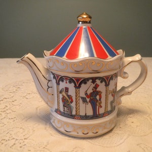 May include: A white porcelain teapot with a red, white and blue striped lid shaped like a circus tent. The teapot features a gold trim and a scene of a band playing music.