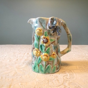 May include: A ceramic pitcher with a floral design featuring yellow flowers and a butterfly. The pitcher has a textured surface and a handle.