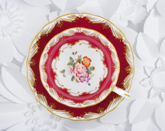 May include: A porcelain teacup and saucer set featuring a deep red border with gold filigree accents. The center of the cup and the saucer display a white background with a hand-painted floral arrangement in shades of pink, orange, and purple.