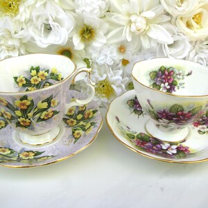 May include: Two vintage teacups and saucers. One teacup is lavender with yellow floral designs and gold trim. The other teacup is white with purple floral designs and gold trim. Both are displayed in front of a white floral background.