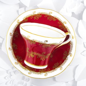 May include: A porcelain teacup and saucer set featuring a deep red background with intricate gold filigree patterns. The cup has a white interior and a gold-trimmed rim and handle, while the saucer displays a matching red and gold design.