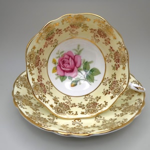 May include: A vintage teacup and saucer set. The cup and saucer are pale yellow with a floral pattern and gold trim. A pink rose is centered in the cup. The set is likely porcelain.