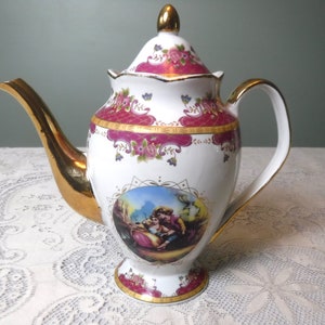 May include: A white porcelain teapot with a gold-trimmed spout and handle. The teapot is decorated with a floral pattern and a scene of two people in a landscape. The lid is also decorated with a floral pattern.