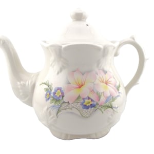 May include: A white ceramic teapot featuring a floral pattern with large pink and yellow blossoms and smaller blue flowers. The teapot has a sculpted, scalloped rim, a curved handle, a spout, and a removable lid with a small knob handle.