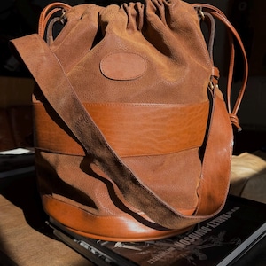 May include: A brown leather bucket bag with a drawstring closure and a long shoulder strap. The bag features a lighter brown band around the middle and a small oval patch. The bag is sitting on top of a book.