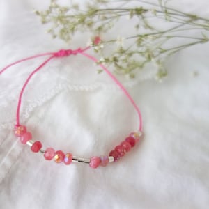 Puede incluir: Un brazalete de cuerda rosa con cuentas rosas y plateadas. El brazalete está atado con un nudo y está sobre una superficie blanca.