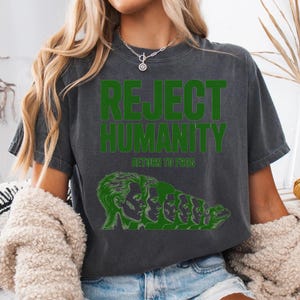 Puede incluir: Camiseta gris oscuro con el texto verde "REJECT HUMANITY" sobre "RETURN TO FROG". Un gráfico verde representa a una persona transformándose en una rana. La camiseta está hecha de un material suave.