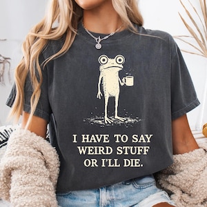 Puede incluir: Una camiseta de manga corta gris carbón con un gráfico color crema de una rana sosteniendo una taza. Debajo de la rana, el texto dice: I HAVE TO SAY WEIRD STUFF OR I'LL DIE. La camiseta tiene cuello redondo y un aspecto desgastado.