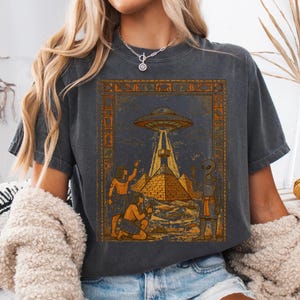 May include: Dark gray t-shirt featuring a vintage-style graphic of a UFO beaming light onto a pyramid, with figures in ancient Egyptian attire. The design includes hieroglyphs and a retro aesthetic, creating a unique and eye-catching design.
