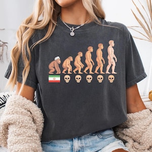 Puede incluir: Camiseta gris oscuro con un gráfico de la evolución humana, con una bandera y calaveras. El diseño muestra una progresión de simio a humano. Una prenda cómoda e informal.