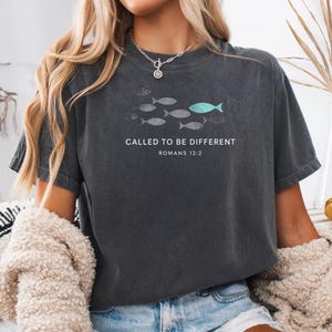 以下が含まれることがあります： 「CALLED TO BE DIFFERENT」と「ROMANS 12:2」の文字が入った濃いグレーのTシャツ。グレーの魚の群れと、ターコイズ色の魚が1匹描かれています。カジュアルで着心地の良いスタイルです。